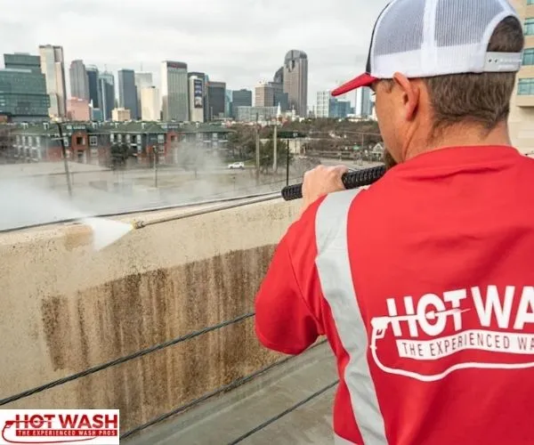 Hot Wash technician wearing a red uniform shirt and white cap uses a pressure washer on a concrete wall overlooking a city skyline.