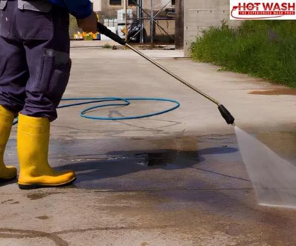 worker in yellow boots spraying a high-pressure water wand to clean a outdoor concrete floor, representing power washing