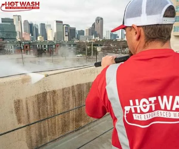 view of a worker in a red Hot Wash shirt pressure washing a dirty concrete ledge overlooking a city skyline, representing power washing