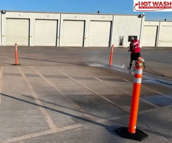 A worker in a red shirt pressure washing a large commercial concrete lot with garage bays in the background