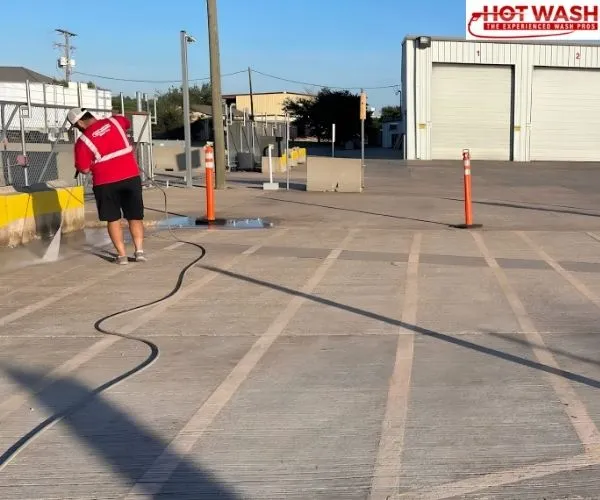 A worker in a red shirt pressure washing a large commercial concrete lot with garage bays in the background